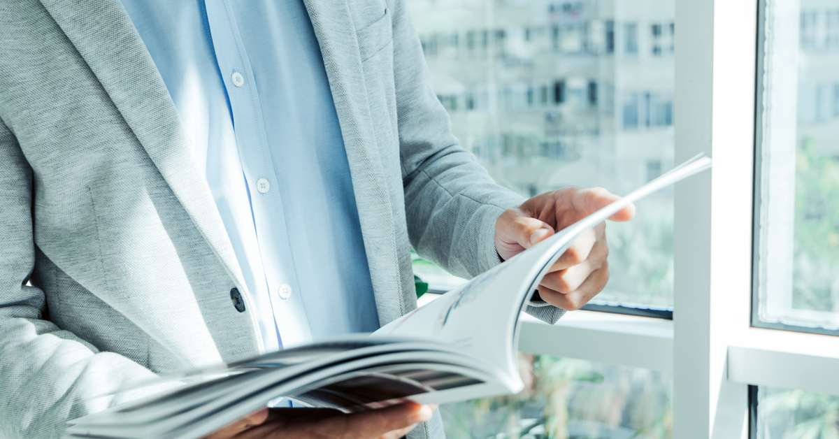 A professional wearing a gray suit stands in front of a large window while looking at a booklet they're holding.