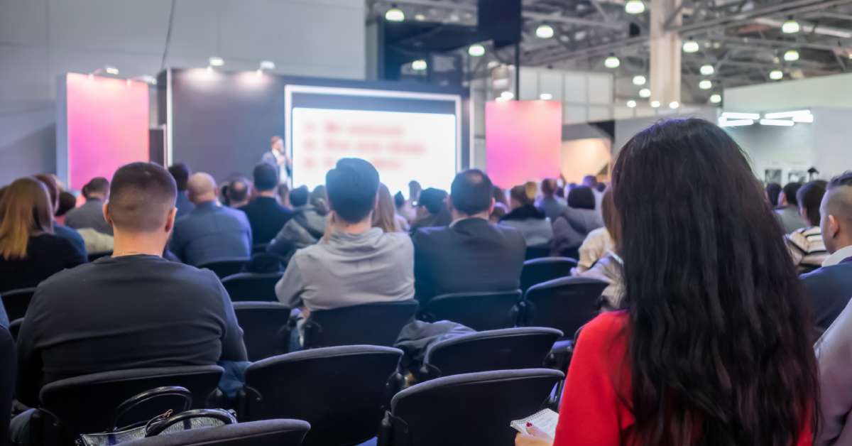 A large crowd of trade show attendees watch a presentation on a stage that has two vibrant banners and a digital screen.