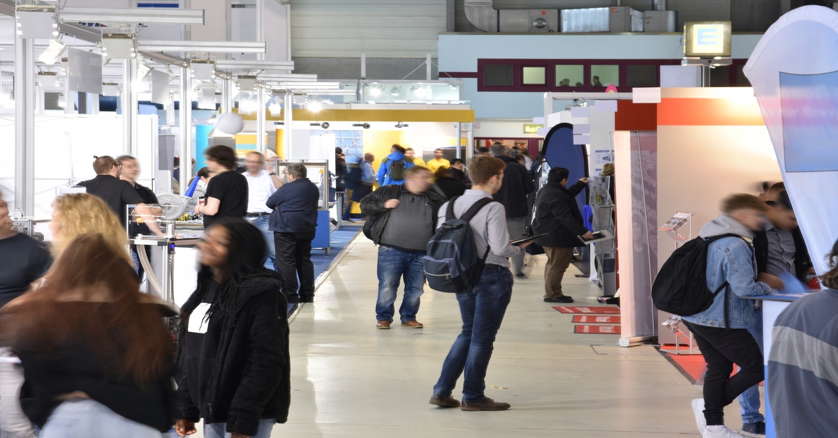People standing in multiple groups at different booths set up at a trade show exhibition in a large facility.