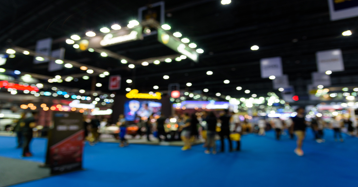 A blurred view of the entrance to a trade show exhibition showcasing multiple booths. Bright lights hang from the ceiling.