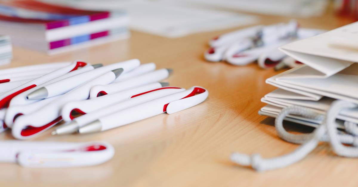 A large pile of white-and-red-colored promotional business pens on a table. A group of bags sits near the pens.