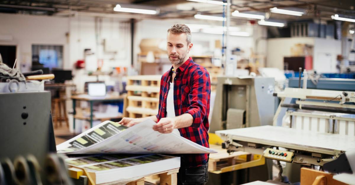A man wearing a flannel in a printing press office unravels a newly printed business banner near a printing machine.