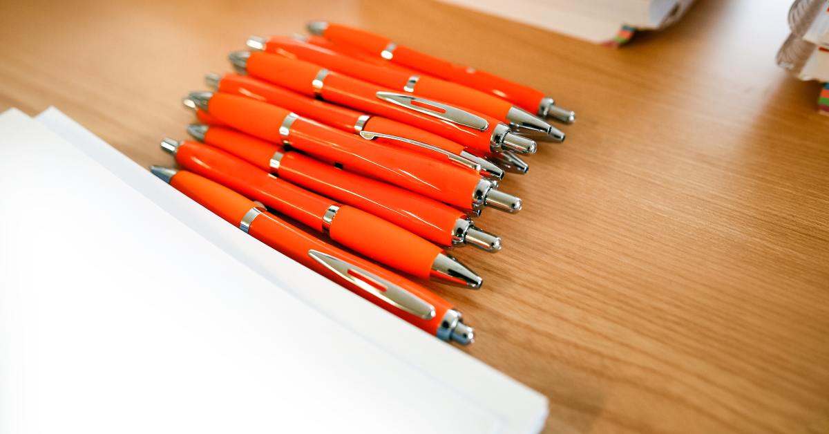 A small pile of orange-colored pens lays on top of a wooden desk near stacks of notebooks and papers.