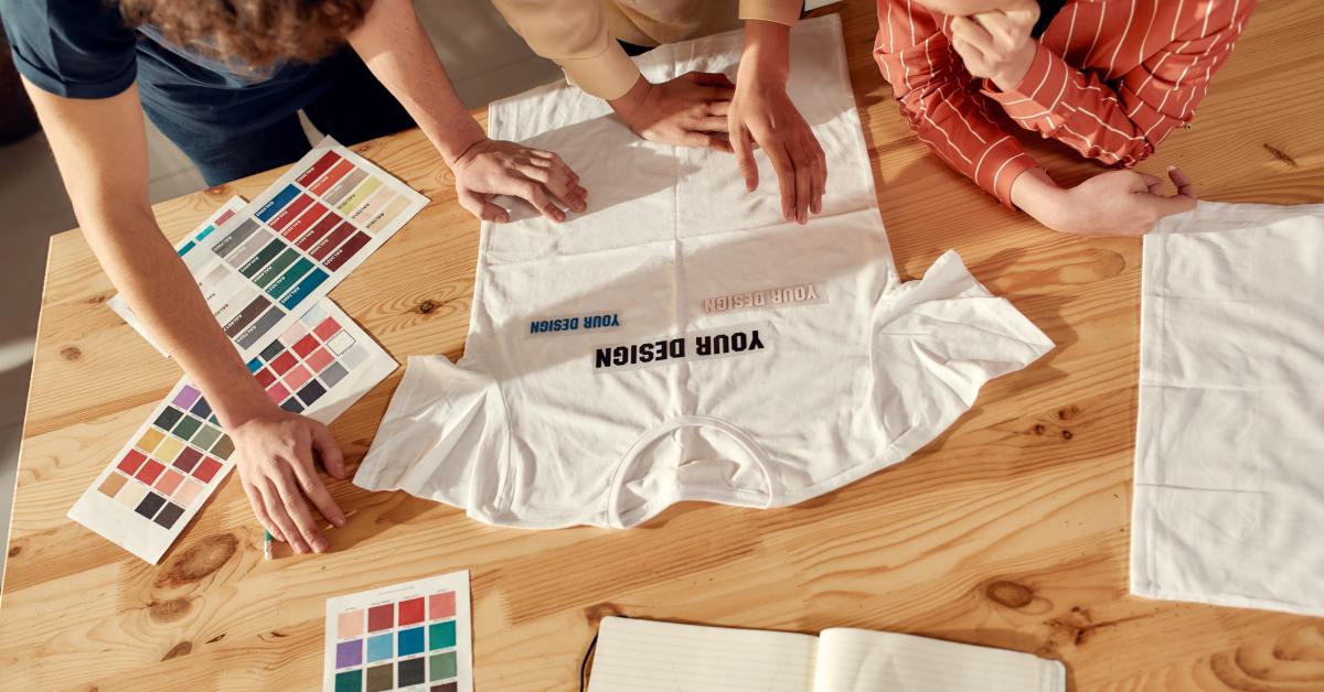 Three people stand around a wooden table and look at a white T-shirt. Multiple color swatches lay nearby.