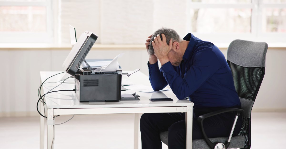 A man sitting in front of a jammed office printer looks down in frustration and puts his hands on his forehead.