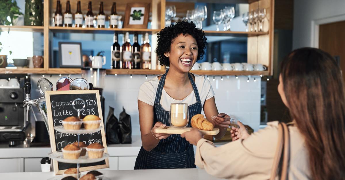 A smiling barista hands a latte and croissant to a customer in a cozy cafe with shelves of bottles, glasses, and baked goods.