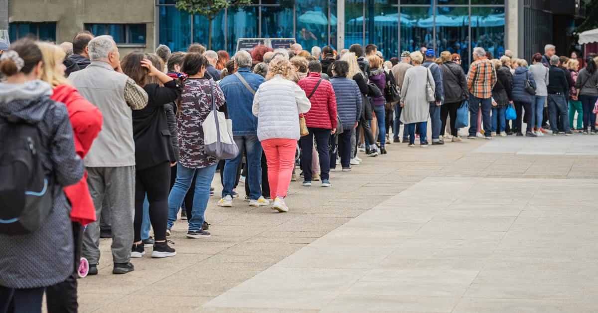A long line of diverse people wait outside of a popular local business. There is municipal building in the background.
