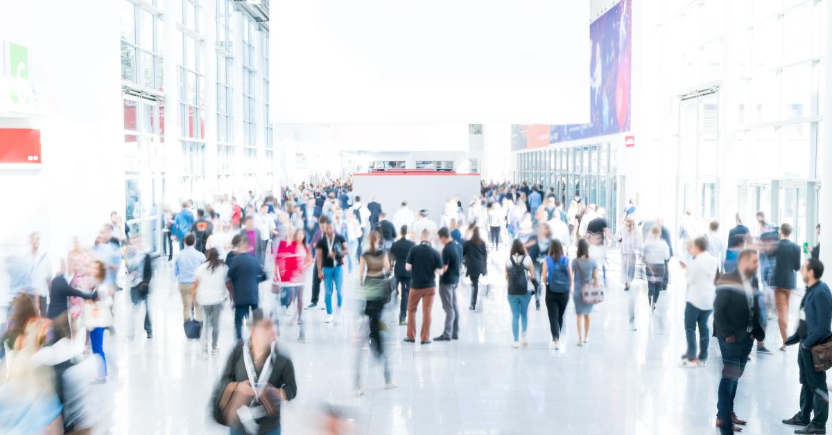 A large, blurred crowd walks through a busy convention center framed by tall glass walls and colorful banners.