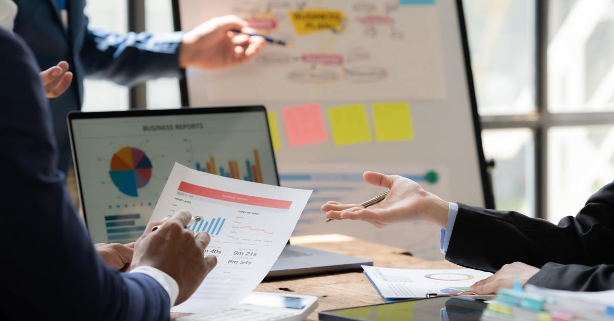A group of people sits at a table full of papers, laptops, and tablets to analyze the data from previous trade show events.