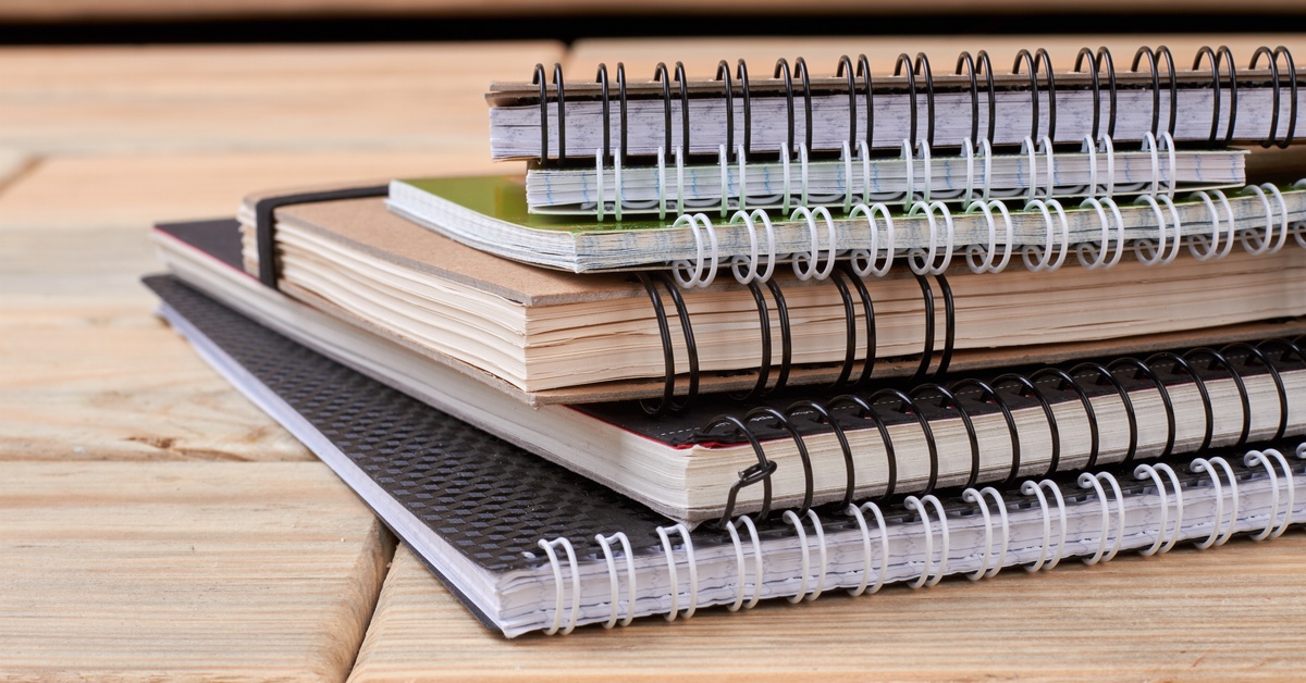 A stack of spiral-bound notebooks in various colors with textured covers and mixed bindings lay on a wooden table.